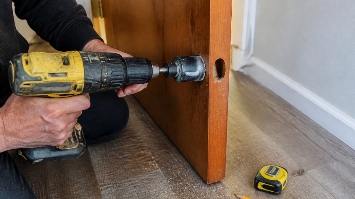 Close-up of a person's hands using a power drill with a hole saw on the edge of a wooden apartment door, with a tape measure and pencil resting on the floor nearby
